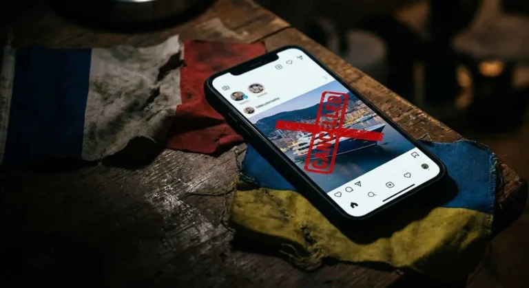 Smartphone mit Social-Media-Post einer Luxusjacht und rotem CANCELLED-Stempel, liegend auf zerknitterten französischen und ukrainischen Flaggen