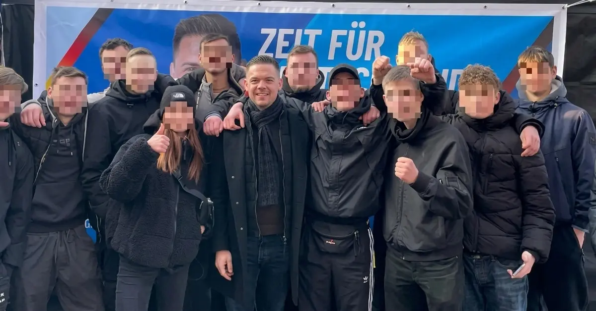 Hannes Gnauck (AfD) posiert grinsend mit einer Gruppe schwarz gekleideter Jugendlicher vor einem AfD-Wahlkampfbanner in Templin